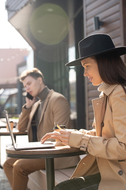 Mulher elegante jovem e contemporânea segurando um cartão de crédito enquanto está sentada à mesa pequena ao ar livre, fazendo compras online e pagando pela compra