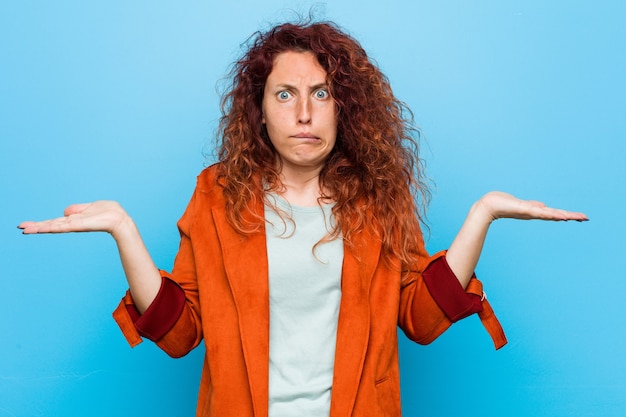 Mulher elegante jovem ruiva confusa e duvidosa, encolhendo os ombros para segurar um espaço de cópia.