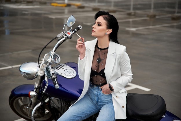 Mulher elegante senta-se em uma motocicleta e fuma um cigarro no estacionamento perto do shopping.