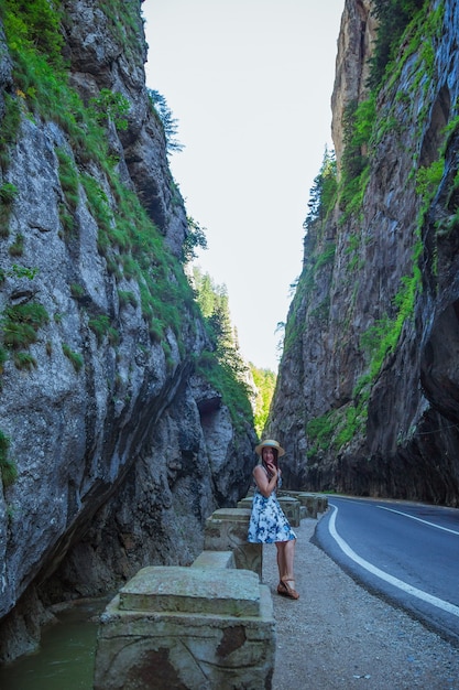 Mulher elegante vestida de vestido azul branco e chapéu de palha, sentada perto do rio da montanha e da estrada perigosa no Canyon de Bicaz. Bicaz Key, maravilhas geológicas da natureza, Transilvânia, Romênia.