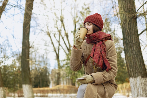mulher em agasalhos quentes bebendo café para viagem de copo de papel enquanto relaxa no banco