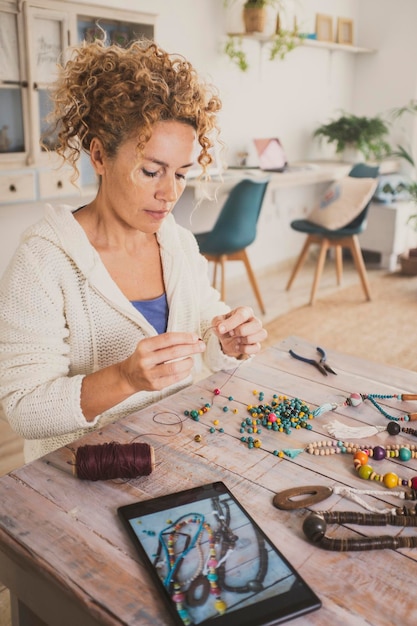 Mulher em casa fazendo joias de miçangas usando instruções on-line em um tablet Trabalho alternativo local de trabalho caseiro Fêmea adulta cria pulseiras e colares como artesã Novos negócios e lazer