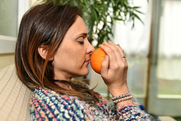 Mulher em casa sentada no sofá cheirando uma laranja com os olhos fechados bom cheiro de fruta dieta saudável