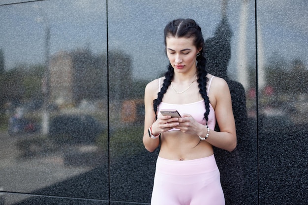 Mulher em forma olhando para a tela do smartphone apoiada contra a parede do edifício