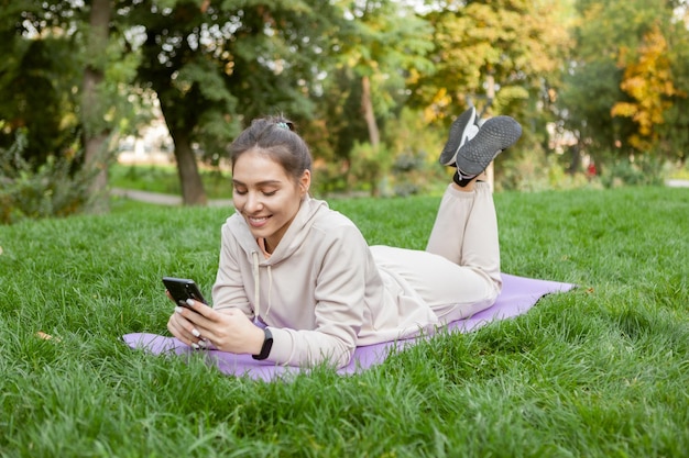 Mulher em forma tem tempo de descanso enquanto está deitada no tapete e usa smartphone no parque Fitness ou ioga ao ar livre