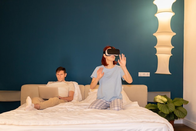 Mulher em óculos VR imersiva no jogo enquanto homem com laptop trabalho na cama diferentes passar tempo em casa