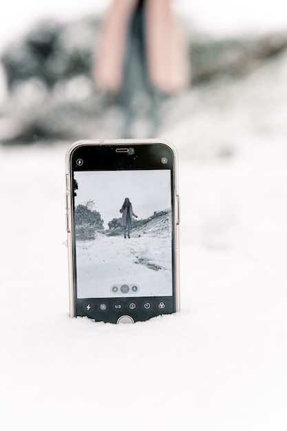 Mulher em pé na floresta de inverno e tirando selfie de smartphone colocado em um monte de neve