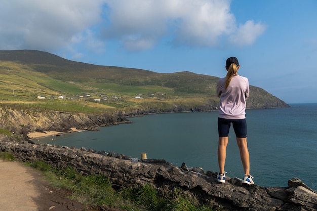 Mulher em pé na parede de pedra, olhando para as falésias na península de Dingle, Irlanda