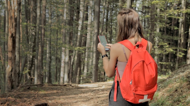 Mulher em pé no meio da floresta, usando o aplicativo de navegação no smartphone, seguindo a rota com ajuda do mapa, olhando para a tela do dispositivo, usando a navegação do smartphone enquanto se perde na floresta.