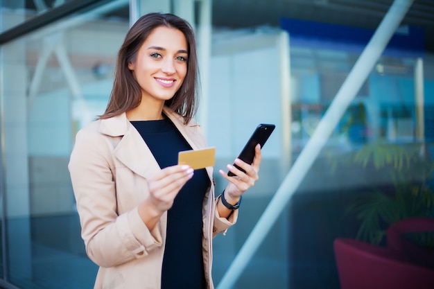 Mulher em pé no saguão do aeroporto ele está usando cartão de crédito ouro e celular para pagar