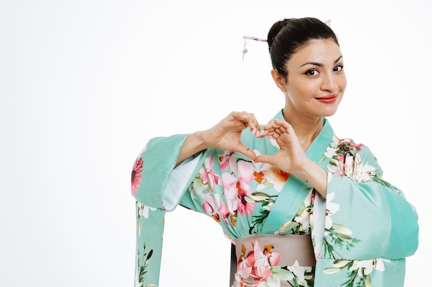 Mulher em quimono japonês tradicional sorrindo fazendo um gesto de coração com os dedos felizes e positivos no branco