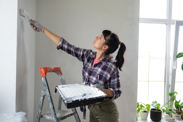 Mulher em rolo de pintura e tinta branca pinta a parede da casa na escada Trabalhos de construção e reparos cosméticos na pintura da parede da casa tingindo o trabalho de acabamento com suas próprias mãos