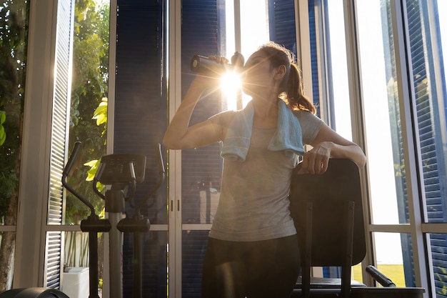 Foto mulher em roupas esportivas, beber água de garrafa no ginásio. há um raio de sol do dia no fundo.