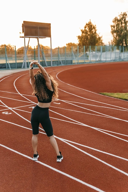 Mulher em roupas esportivas com cabelo encaracolado permanente está se esticando em um estádio pela manhã.