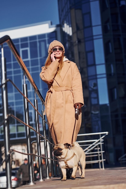 Mulher em roupas quentes passear com seu cachorrinho perto de negócios construindo isso em segundo plano e falando por telefone.