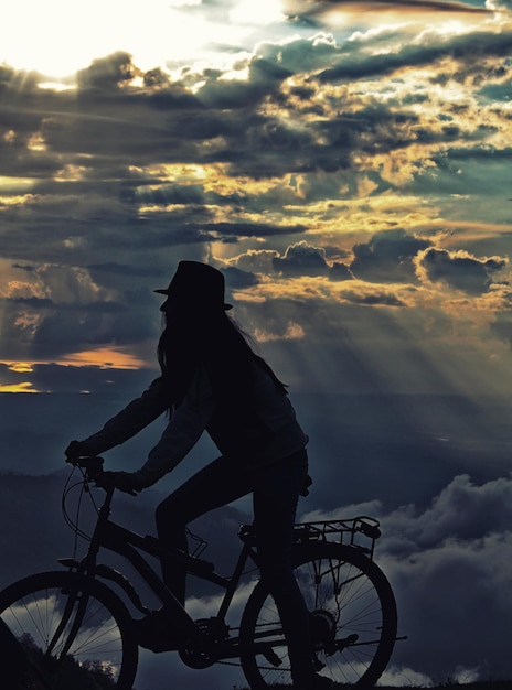 Foto mulher em silhueta andando de bicicleta contra a paisagem nublada durante o pôr do sol
