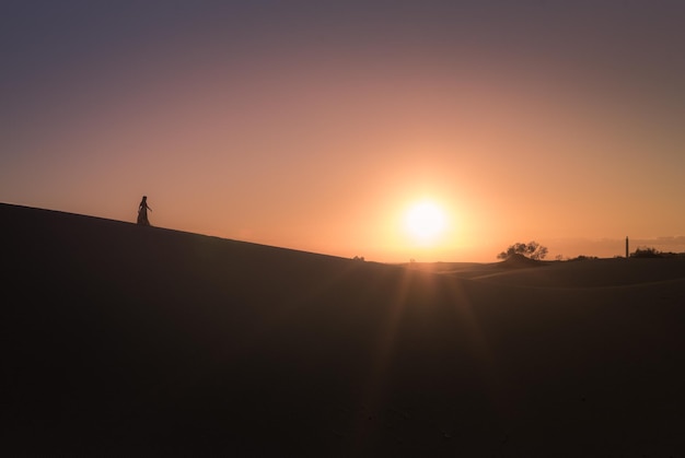 Mulher em silhueta de pé em uma duna de areia contra um céu claro durante o pôr do sol
