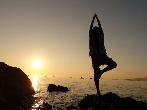 Foto mulher em silhueta fazendo ioga em uma rocha no mar contra o céu durante o pôr do sol