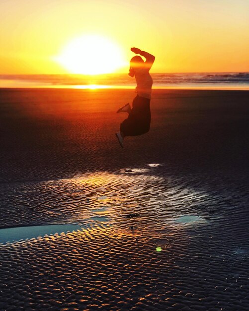 Foto mulher em silhueta pulando na praia contra o céu durante o pôr do sol