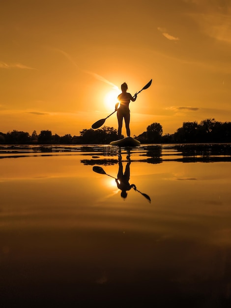 Mulher em sup board paddle boarding no pôr do sol reflexão na água