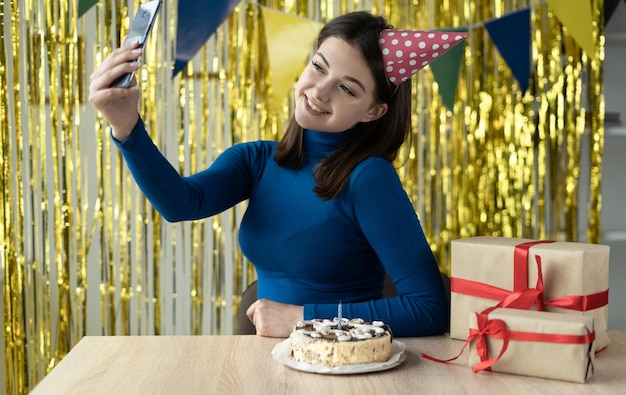 Foto mulher em um boné festivo com um sorriso no rosto senta-se em seu aniversário em uma festa online usando um smartphone e um aplicativo bate-papo por vídeo com amigos à distância