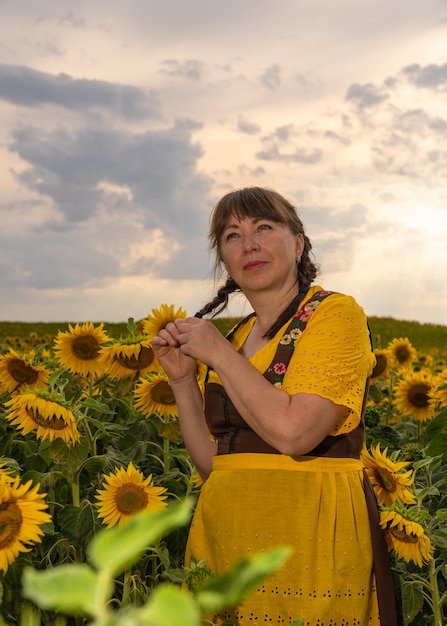 Mulher em um campo de girassóis e ela usa uma blusa amarela um vestido marrom e um avental amarelo