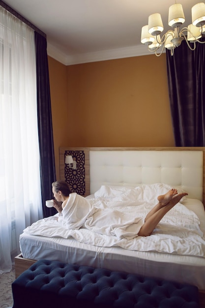 Mulher em um roupão branco está sentada na beira da cama e tomando chá em uma caneca em uma grande janela do hotel