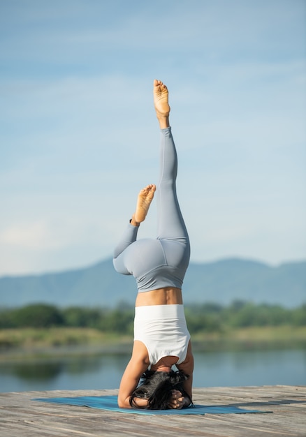 Foto mulher em um tapete de ioga para relaxar no parque. jovem mulher asiática desportiva praticando ioga, fazendo exercícios de headstand, malhando, vestindo roupas esportivas, calças e top.