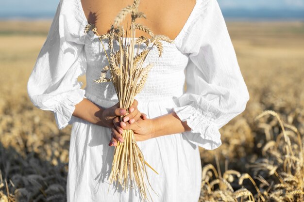 Mulher em um vestido branco fica no campo com uma pessoa de trigo segura em um pacote de mãos de colheitas de espigas maduras ...