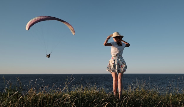 Mulher em uma colina segurando um chapéu nas mãos e olhando para um parapente