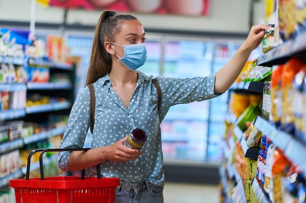 Mulher em uma máscara de proteção médica escolhe e leva produtos alimentares de uma prateleira em uma mercearia