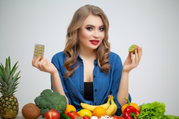 Foto mulher em uma mesa segurando um kiwi e comprimidos em um fundo de frutas e legumes.