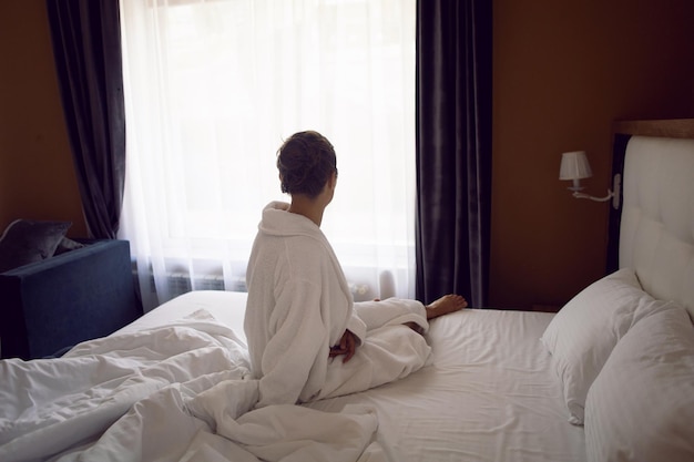 Mulher em uma túnica branca está sentada em uma cama perto da janela em um apartamento de hotel depois de um banho