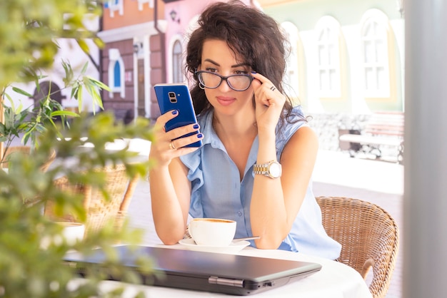 Mulher em vidro trabalhando remotamente com laptop e telefone no café. bela morena usando o notebook no café. feliz empresária chamando no telefone móvel e tendo. freelance