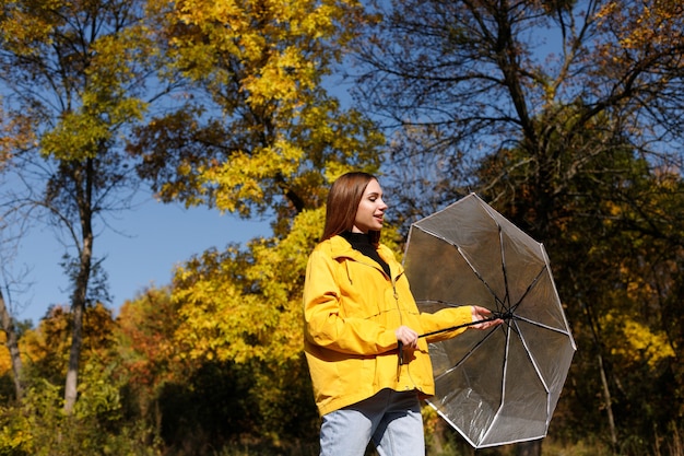 Mulher emocional sorri e desvia o olhar com um guarda-chuva em folhas de outono com um fundo de dia brilhante