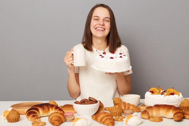 Mulher encantada satisfeita vestindo camiseta branca isolada sobre fundo cinza bebendo chá com bolo sorrindo com expressão feliz desfrutando de pastelaria saborosa