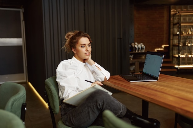 Mulher encantadora com cabelo loiro usando laptop sem fio enquanto trabalha em um café moderno