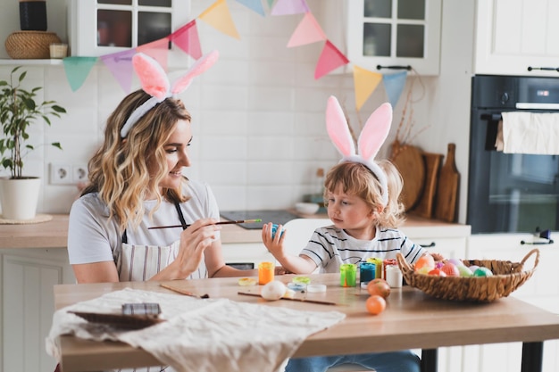 Mulher encantadora e seu filho de dois anos em orelhas de coelho pintam ovos de Páscoa com tintas sentados à mesa na cozinha