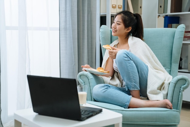 mulher encantadora japonesa asiática tomando café da manhã enquanto trabalhava com laptop laptop na sala de estar em casa no sofá de conforto. bela dama desfruta de pão crocante saboroso no sofá único olhe pela janela