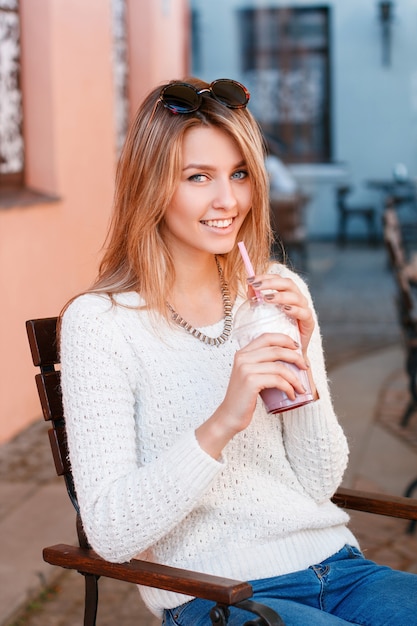 Mulher encantadora jovem hippie feliz com um lindo sorriso em uma camisola de malha em jeans com óculos de sol é vertida em uma cadeira de madeira e bebendo um batido doce. Linda garota descansando.