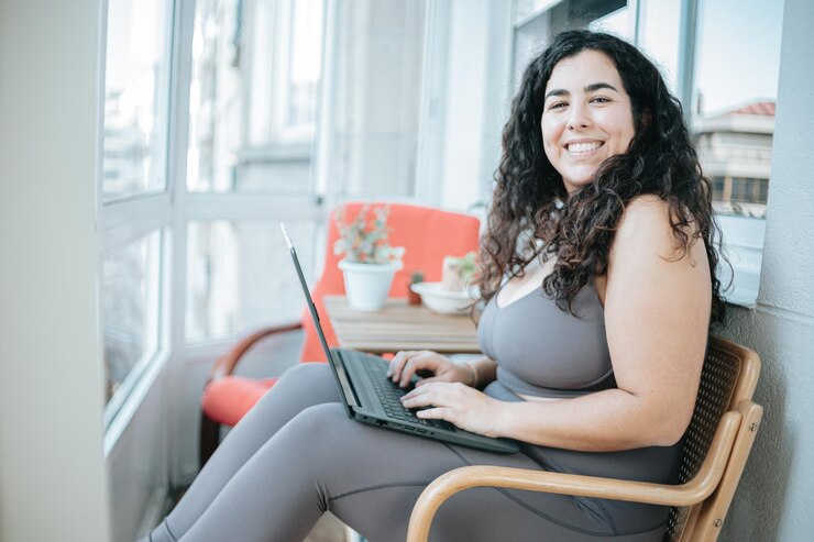 Foto mulher encaracolada feliz plus size sorrindo para a câmera enquanto usa um laptop para preparar a postagem e fazer treinamento on-line roupas de treinamento ficando em forma para o conceito de verão preparação do corpo na praia