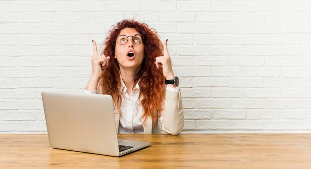 Mulher encaracolada jovem ruiva trabalhando com seu laptop apontando de cabeça com a boca aberta.