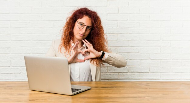 Mulher encaracolada jovem ruiva trabalhando com seu laptop, sorrindo e mostrando uma forma de coração com as mãos.