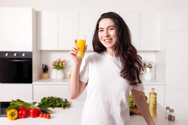 Mulher encaracolada segurando um copo de suco de laranja e sorrindo na cozinha moderna branca