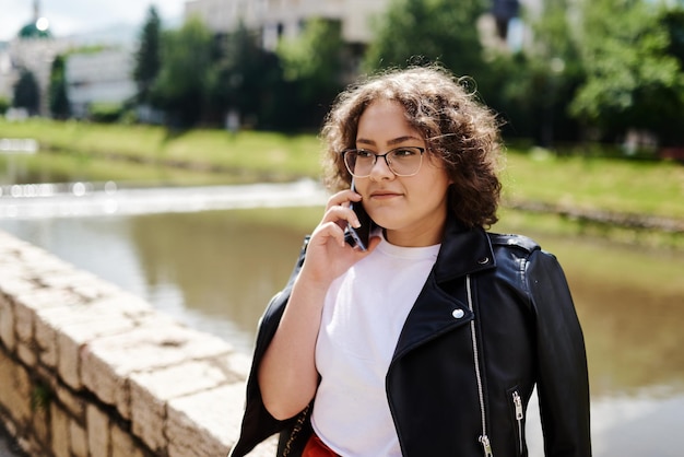 Mulher encaracolada sorridente usando óculos escuros da moda caminha pela rua central da cidade e usa seu telefone