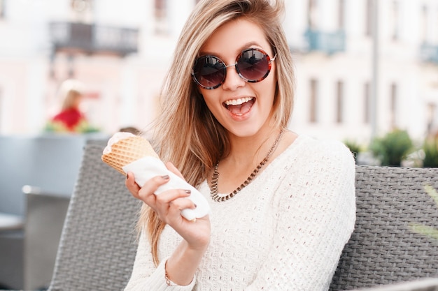Mulher engraçada alegre jovem hippie em um suéter vintage de malha branca com sorvete de baunilha doce em um cone sentado ao ar livre. Garota atraente e alegre.