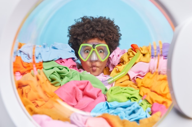 Mulher engraçada com cabelo afro coberto com roupa multicolorida na máquina de lavar fazendo lábios de peixe usar máscara de mergulho fingindo mergulhar de dentro da máquina de lavar.