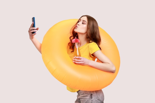 Mulher engraçada com cabelo castanho ondulado em camiseta amarela segurando o anel de borracha laranja e fazendo selfie no telefone faz gesto de beijo descansando no resort Foto de estúdio interior isolada em fundo cinza