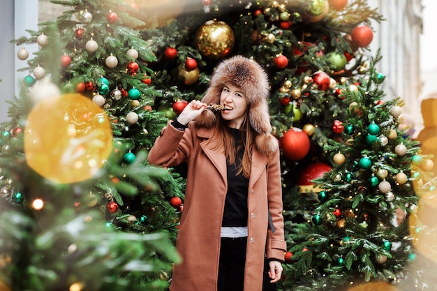 Mulher engraçada com chapéu com abas de orelha está mordendo o pirulito entre as luzes borradas da árvore de Natal na feira de Natal.