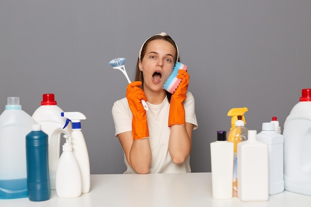 Mulher engraçada e lúdica vestindo camiseta branca e luvas laranja posando isoladas sobre fundo cinza segurando a esponja para limpeza como um telefone mantém a boca aberta se divertindo enquanto faz as tarefas domésticas